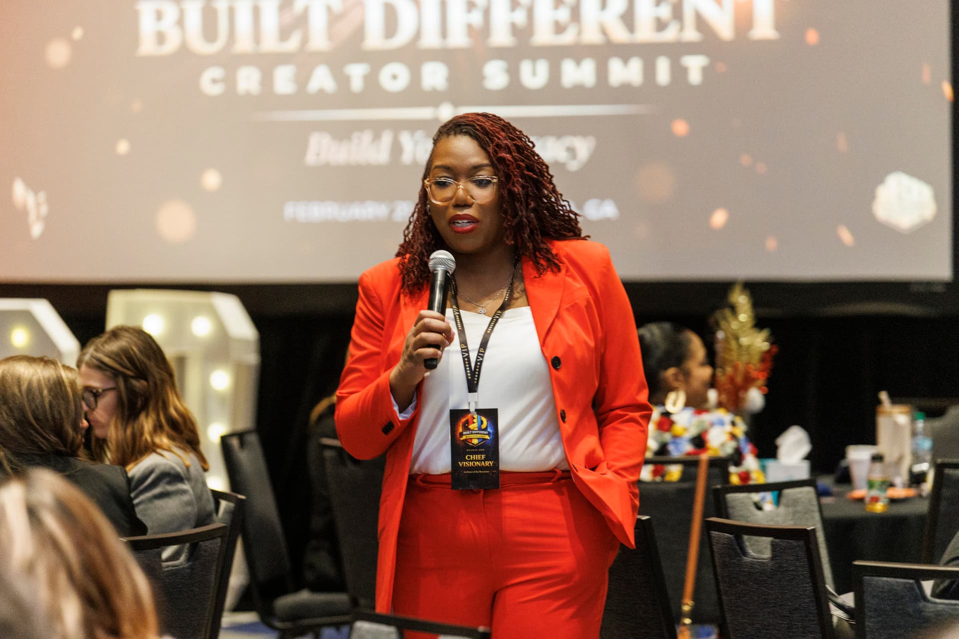 Coach Key in a red suit speaking into a handheld microphone on the floor at the Built Different Creator Summit