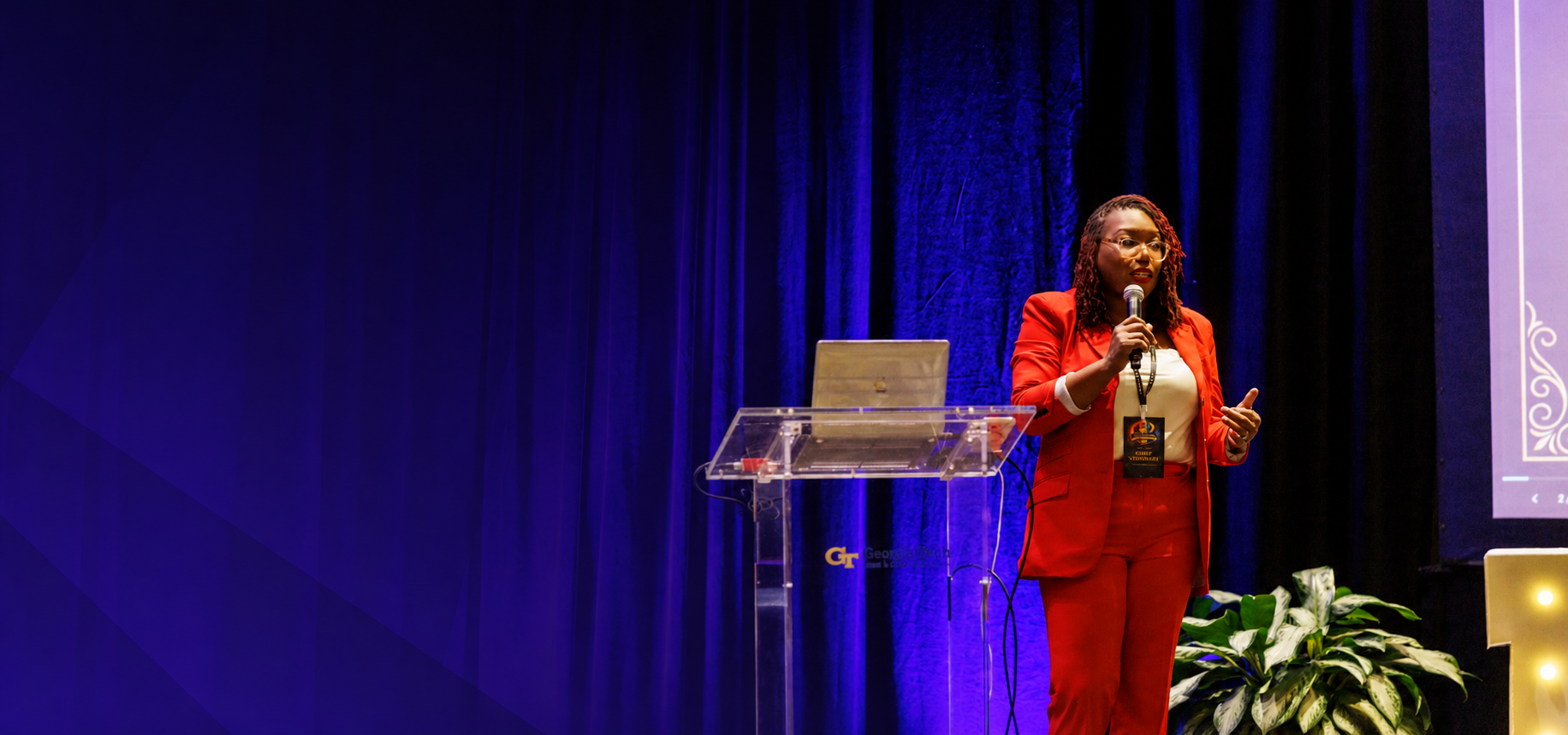 Coach Key, LaKeysha Cobbs-Hayes, speaking into a handheld microphone on stage in a red suit beside a clear lectern